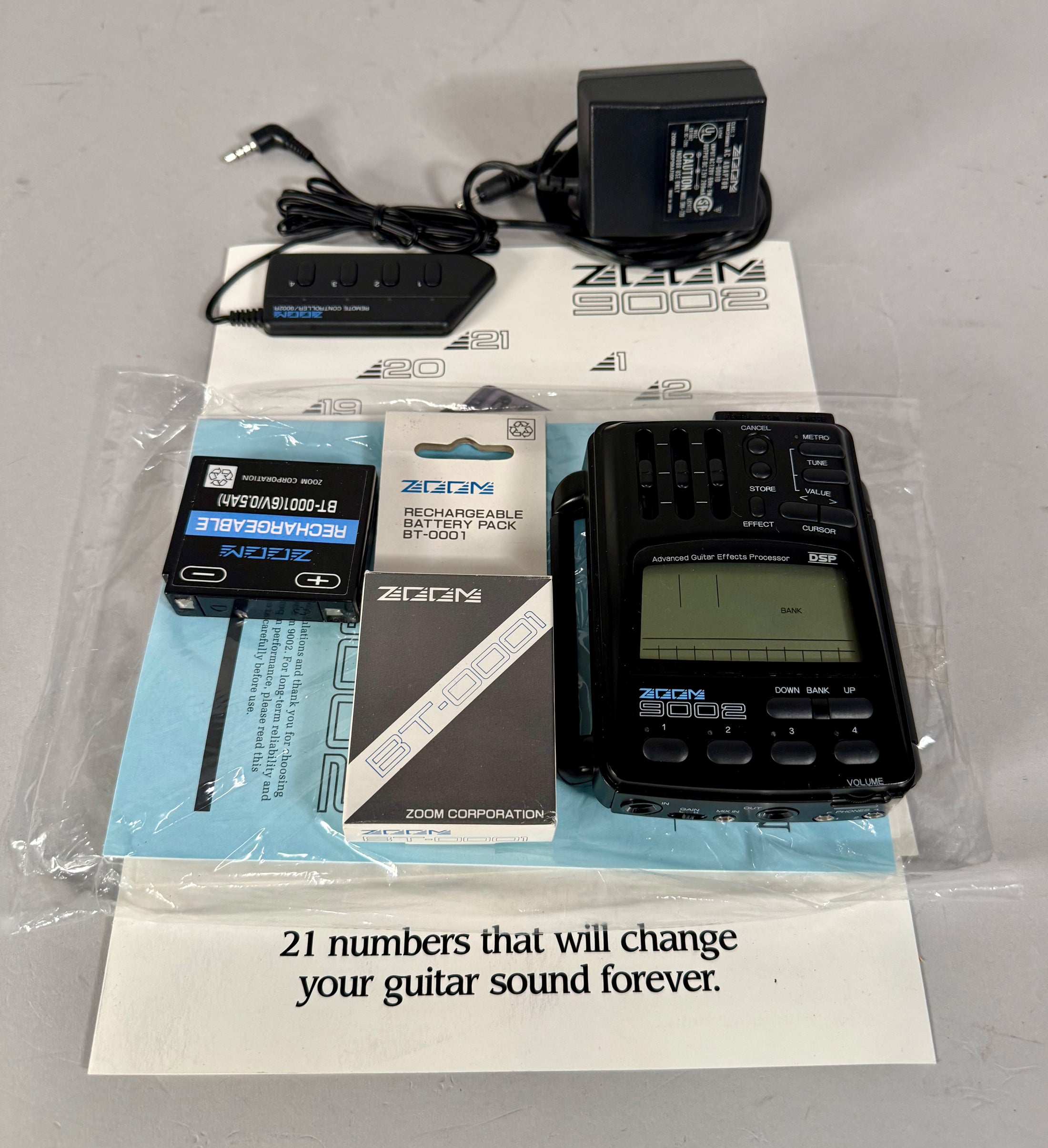 A black handheld effects processor with a screen and sliders, shown with its power adapter, remote, and battery pack.