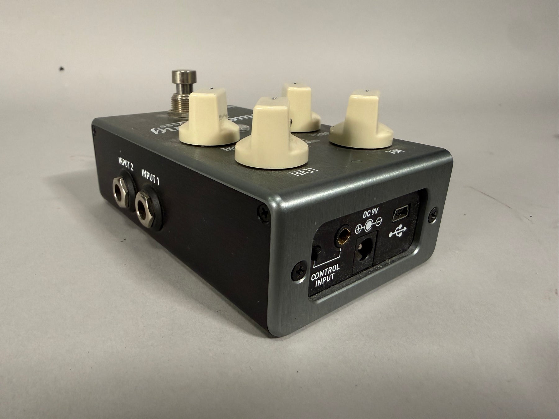Dark gray effects pedal, angled from the left, showing the left side with two input jacks and the rear panel with power and USB ports.