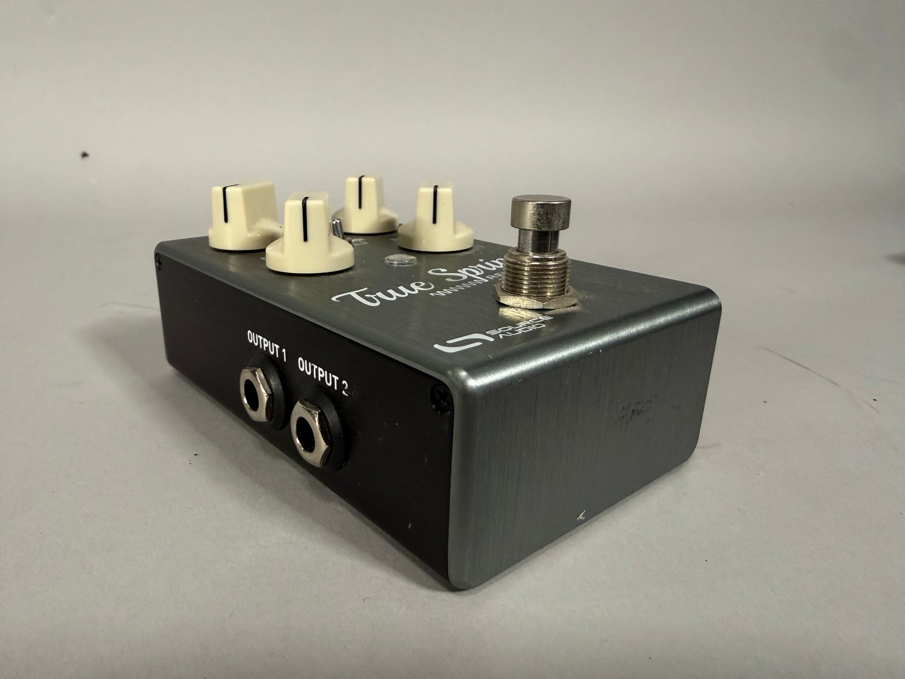 A dark gray effects pedal with a brushed metal top, four cream-colored knobs, a silver footswitch, and two output jacks.