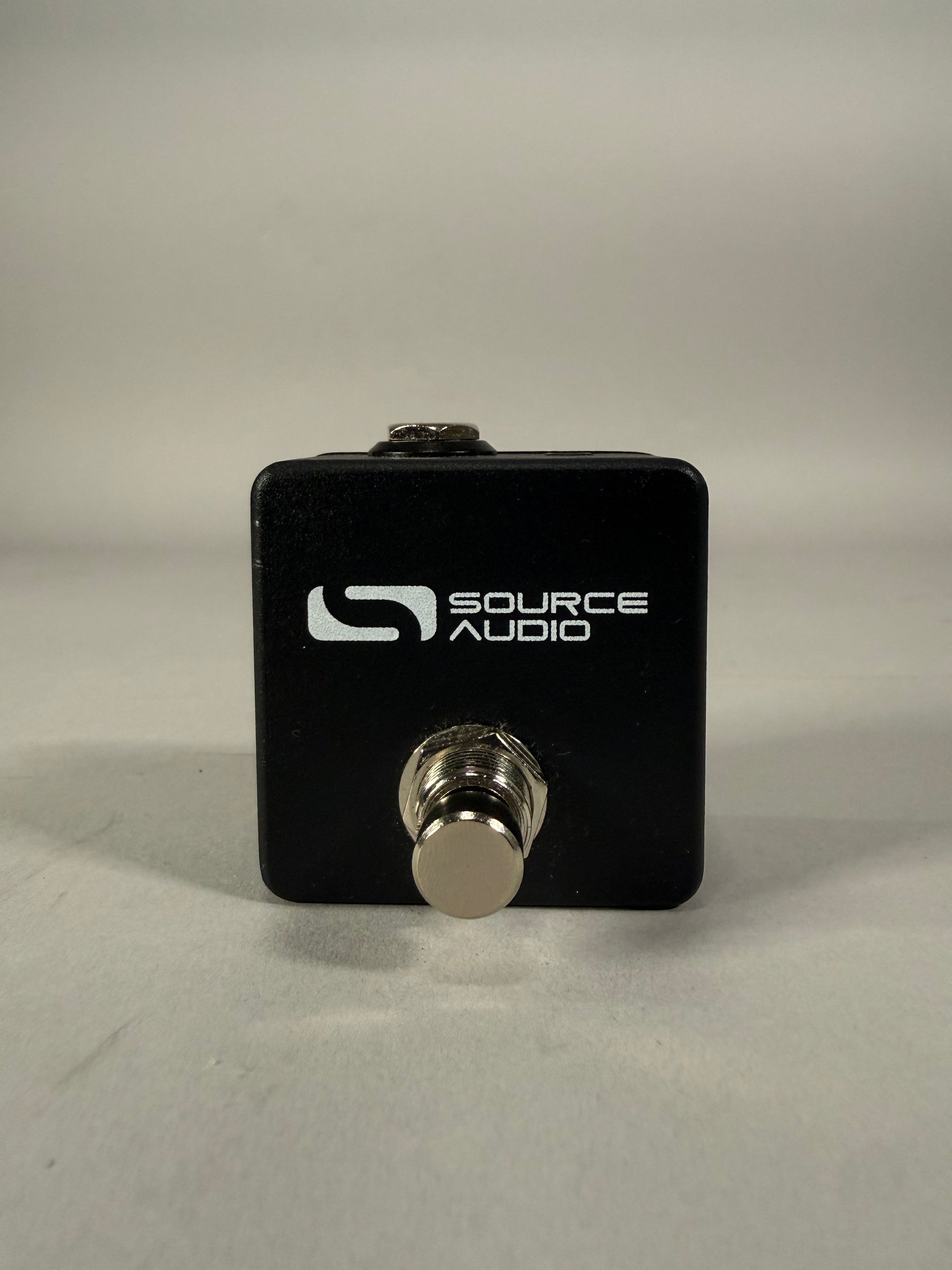 A black, cube-shaped tap switch pedal with a single chrome footswitch on the front and a small chrome jack on top.