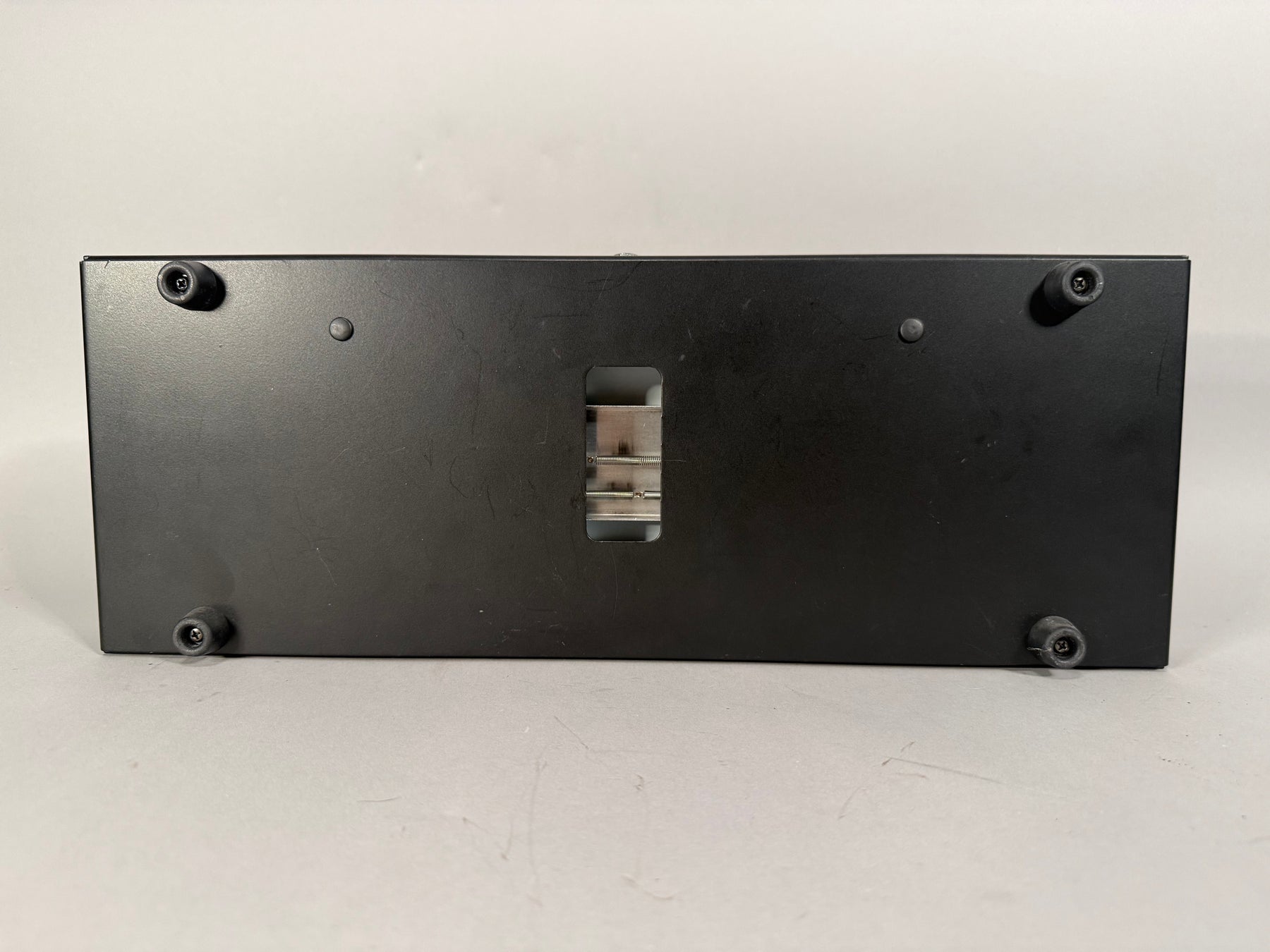 The black metal enclosure is viewed from the bottom, showing four rubber feet and a central rectangular opening with internal metal springs.
