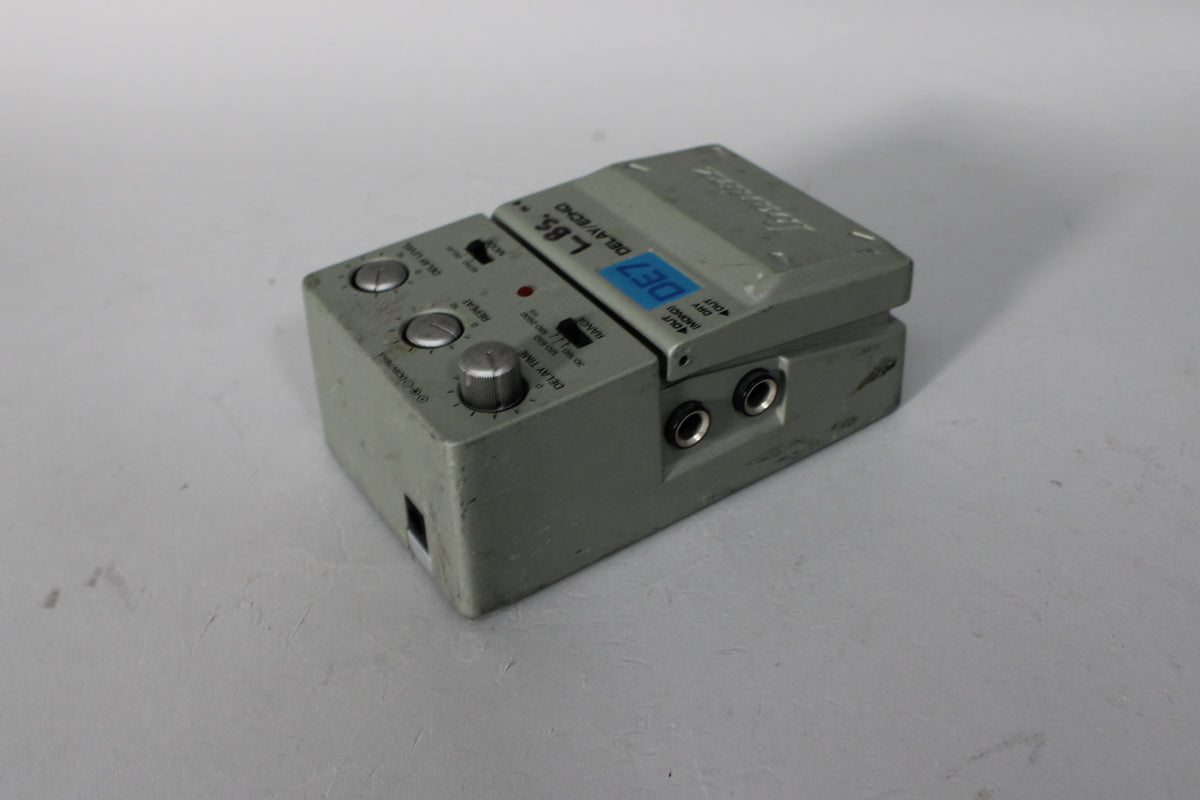 A gray pedal with three silver knobs, two switches, and two input jacks, viewed from the front-right.