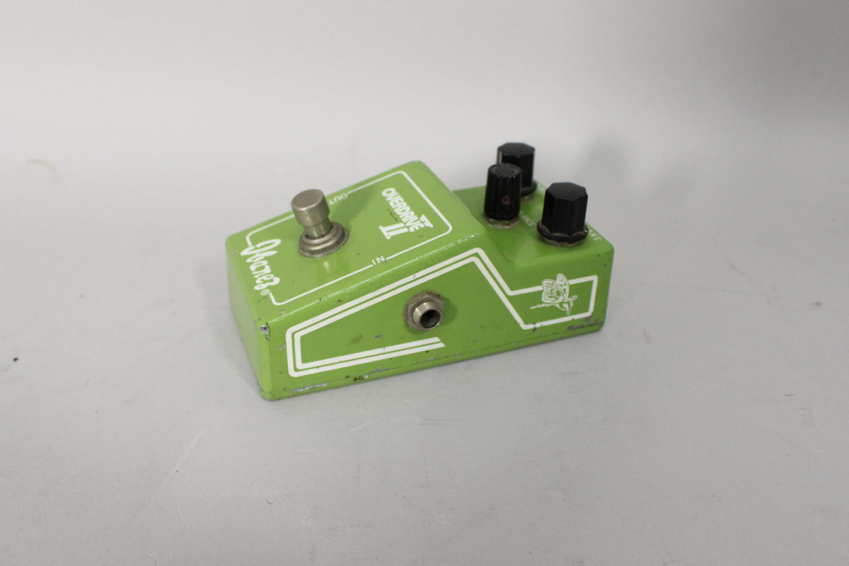 A worn green overdrive pedal, angled from the front-right, showing three black knobs, a silver footswitch, and an input jack.