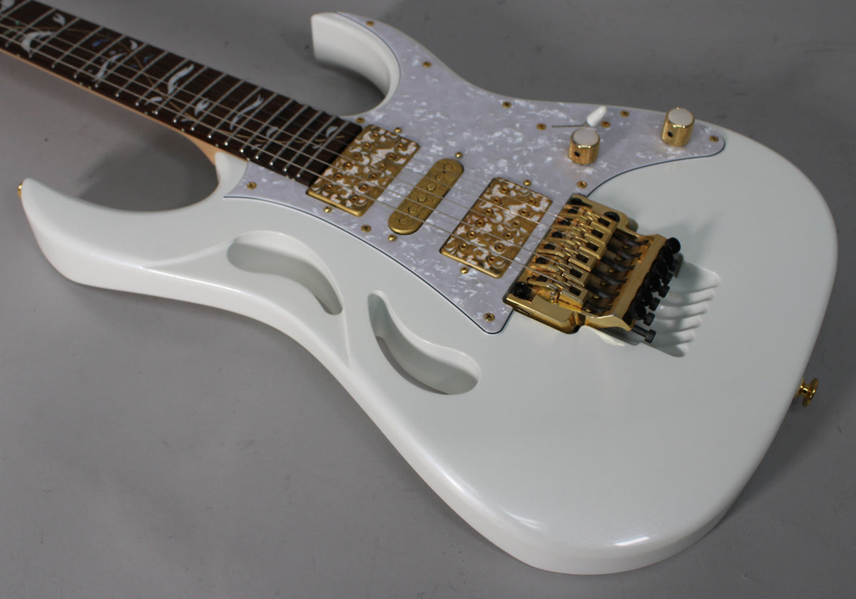 The white electric guitar is angled from the neck, showing its pearloid pickguard, gold hardware, and three carved-out "monkey grip" holes.
