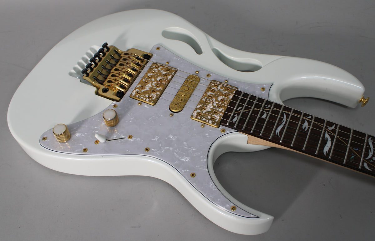 White electric guitar, angled from above, showing the body, gold hardware, pearloid pickguard, and ornate fretboard inlay.
