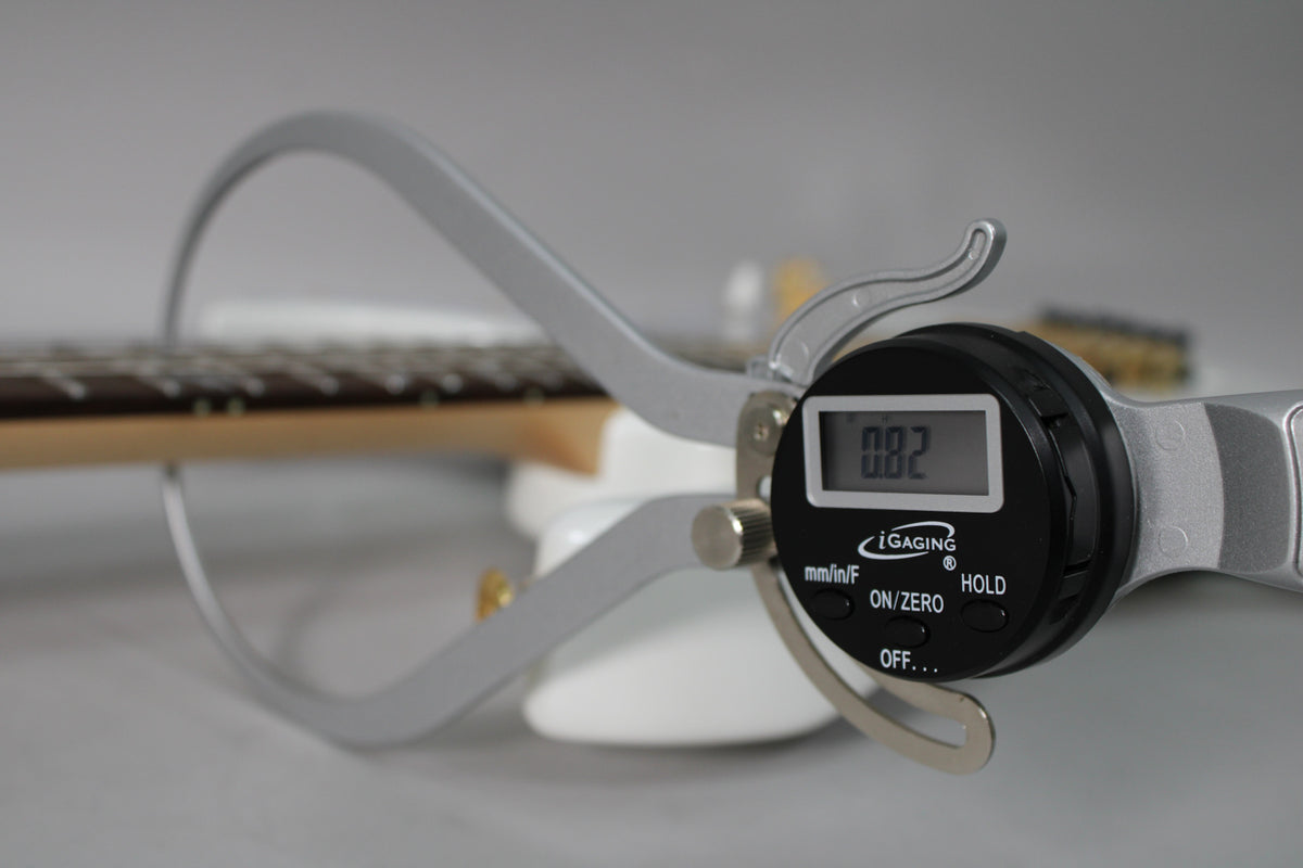 A digital caliper measures the guitar's fretboard and strings, showing "0.02" on its screen, with the white body blurred in the background.