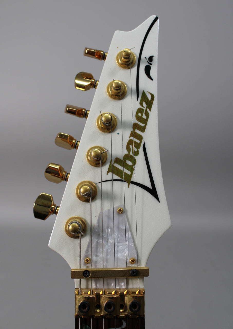 Close-up of the white headstock with gold tuners, a gold locking nut, and a pearloid truss rod cover.