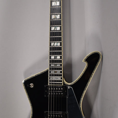 Full-length, angled view of a glossy black electric guitar with a unique body shape, chrome hardware, and pearloid block inlays.