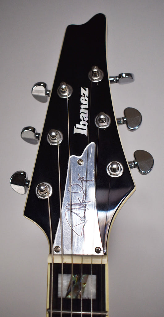 The black gloss headstock with white binding features chrome tuners, a silver truss rod cover with a signature, and block inlays.