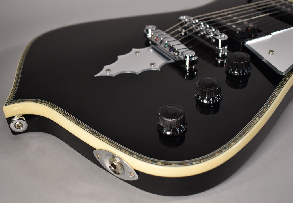 The glossy black guitar body, framed from the lower bout, features cream binding, three black control knobs, and a chrome bat-wing tailpiece.