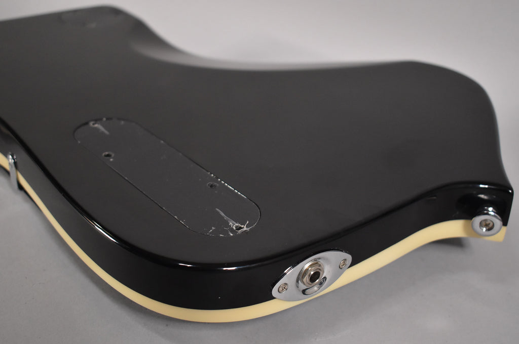 The glossy black guitar body's back shows a control cavity cover with scratches, a chrome output jack, and cream binding.