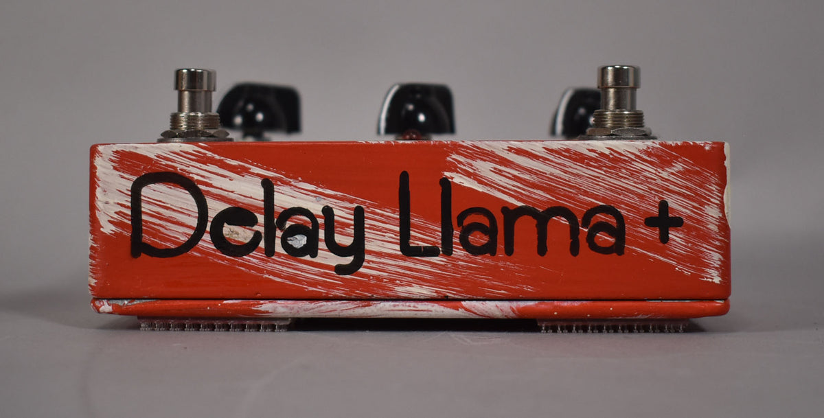 A red guitar pedal with white brushstrokes and "Delay Llama +" in black text, with three knobs and two footswitches visible.