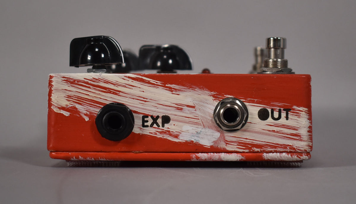 Side view of a distressed red pedal with white brushstrokes, showing "EXP" and "OUT" jacks, and black control knobs on top.