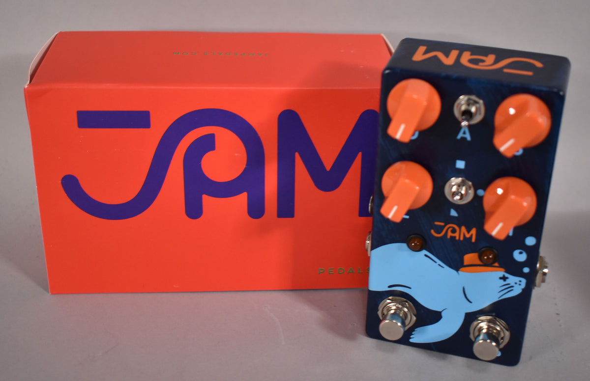 A dark blue pedal with a light blue seal wearing a hat, four orange knobs, two toggle switches, and two footswitches, next to its orange box.