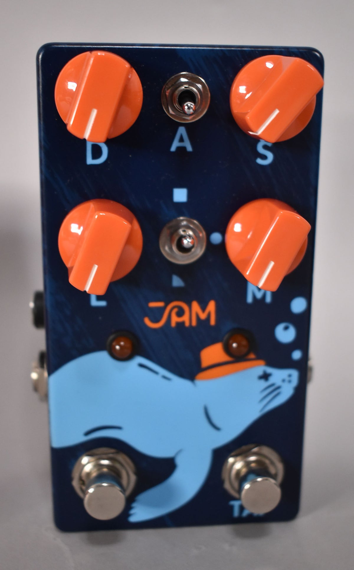 A dark blue pedal with orange knobs and switches, featuring a light blue cartoon seal wearing an orange hat.