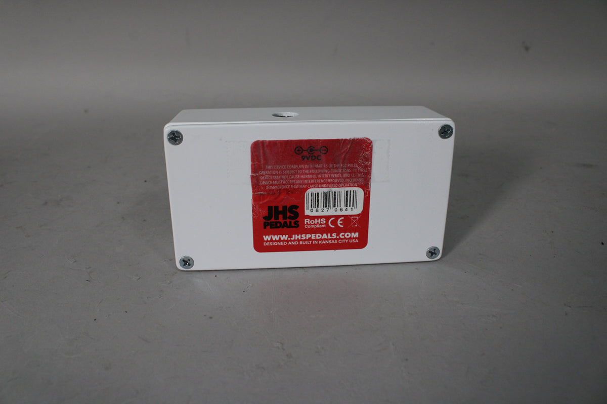 The white pedal is viewed from the back, showing a red label with a barcode and 9VDC power input, secured by four screws.