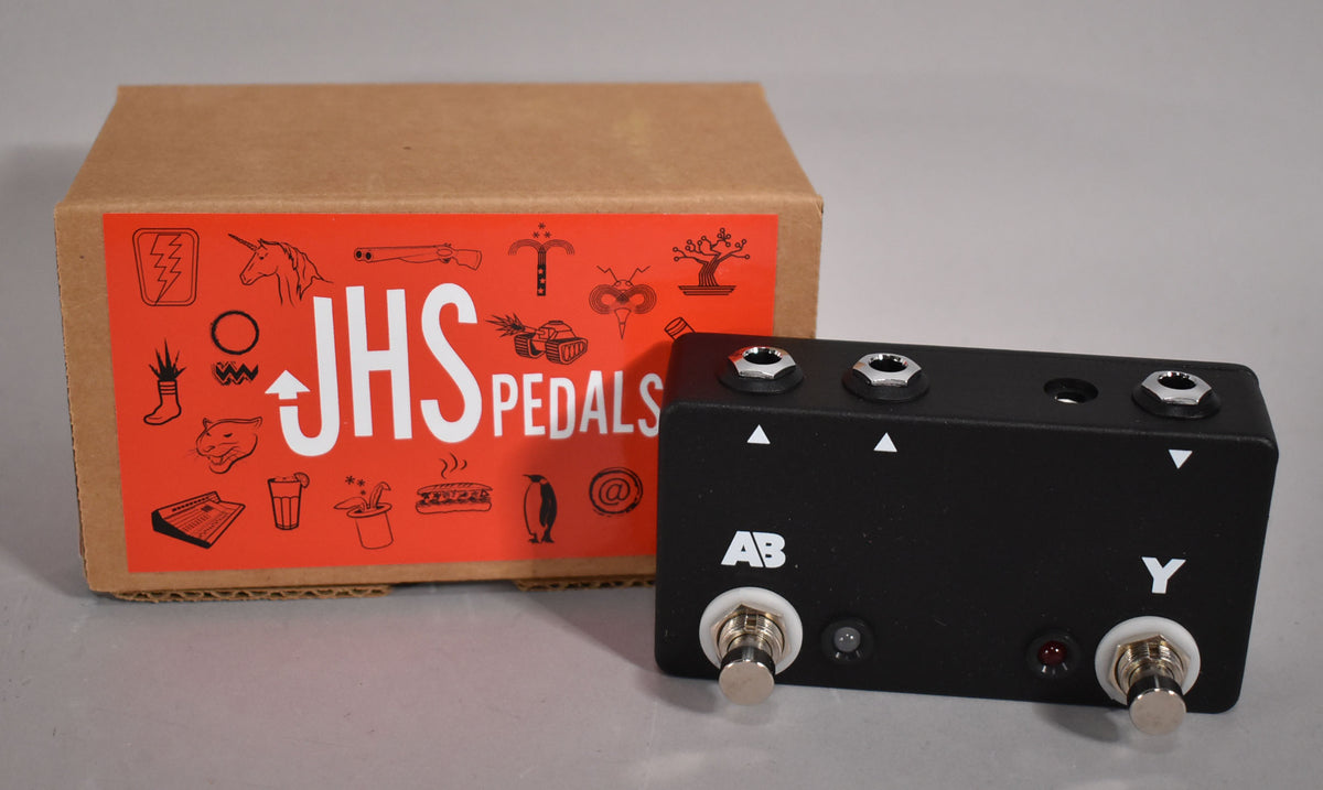 A black metal pedal with two footswitches, two indicator lights, and three input jacks, next to a brown cardboard box.
