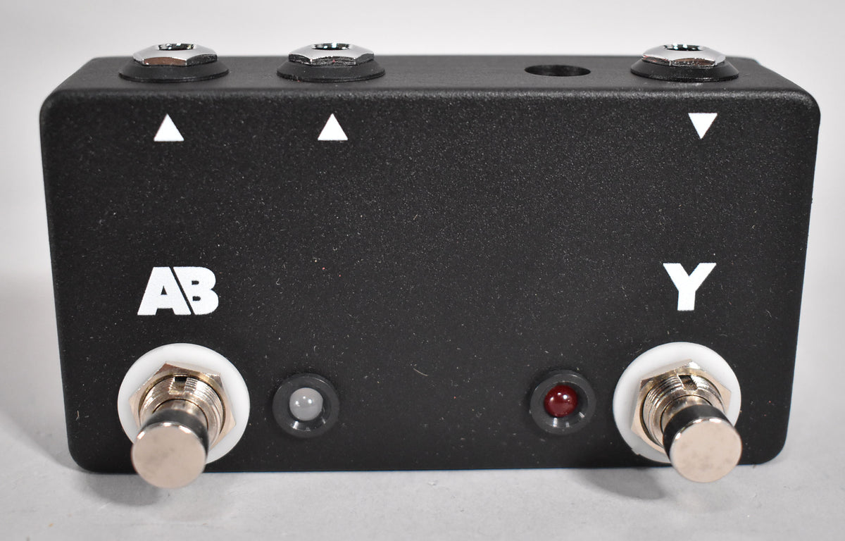 A black, textured pedal with two footswitches, two indicator lights (one gray, one red), and three input jacks on top.
