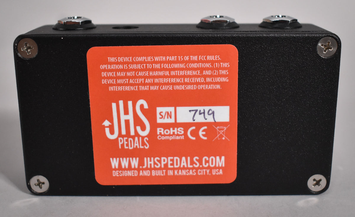 The black pedal's top edge is visible, showing three chrome input jacks and a large orange sticker with product information.