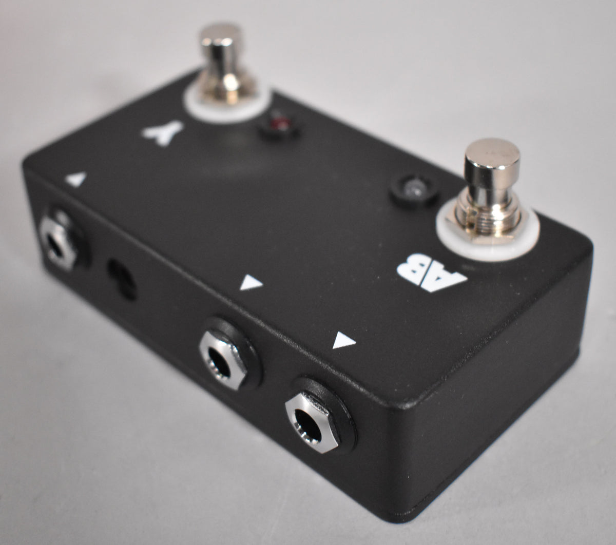 A black pedal with two chrome footswitches, three chrome input jacks, and a red LED, viewed from the front-right.