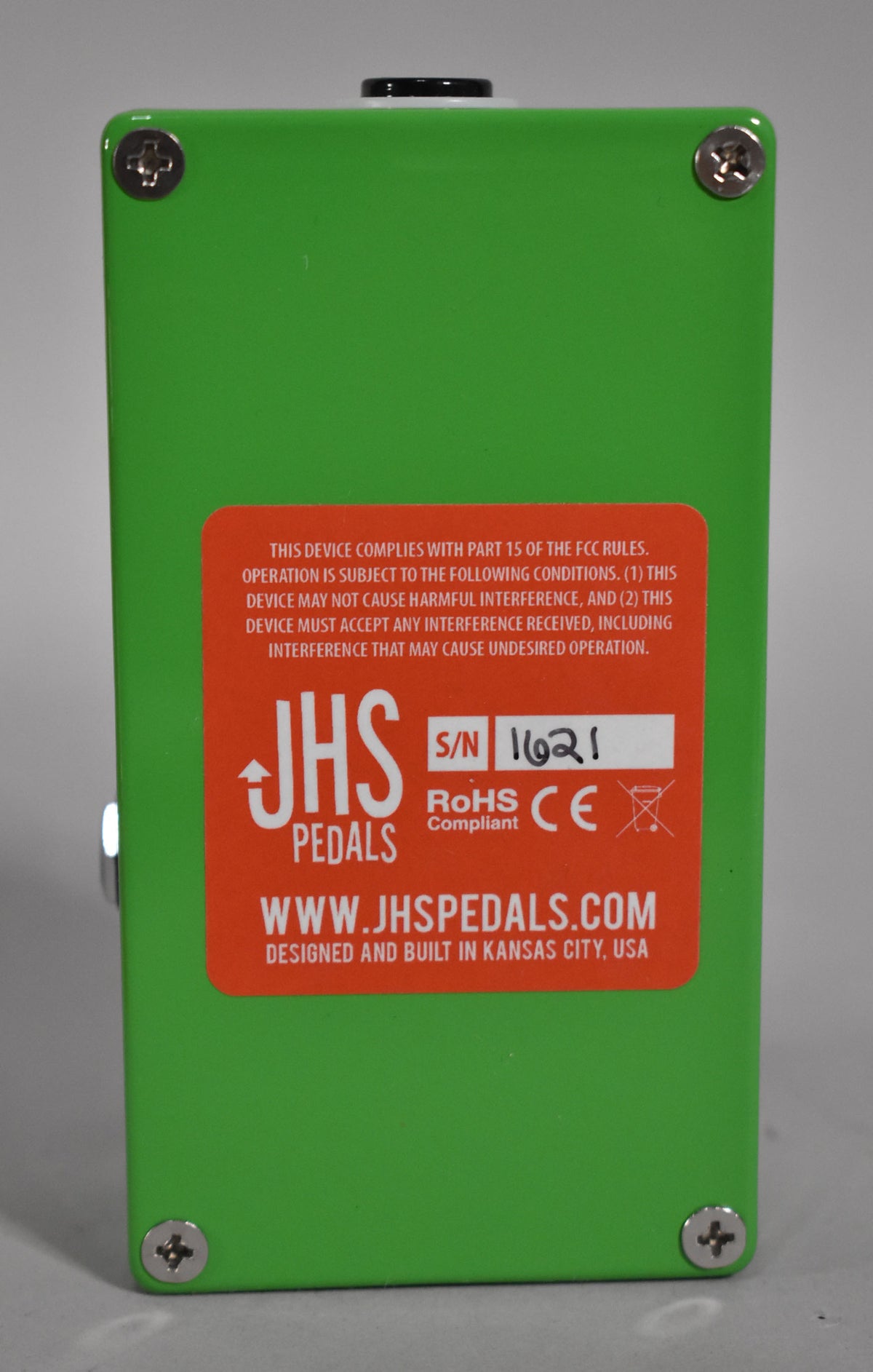 The bright green pedal is viewed from the back, revealing a large orange sticker with white text and a handwritten serial number.
