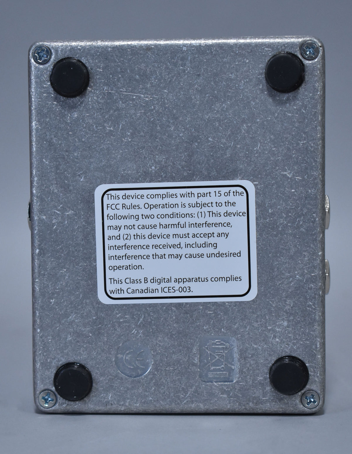 The pedal's textured silver bottom plate is shown, with four black rubber feet and a white regulatory sticker centered.