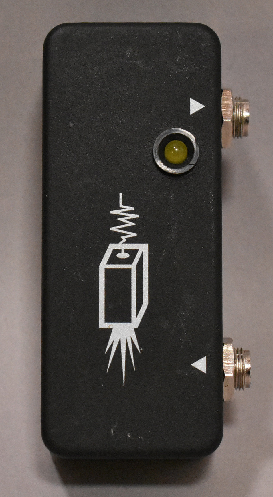 Top-down view of a rectangular, textured matte black pedal with a yellow LED, two input jacks, and a white rocket graphic.