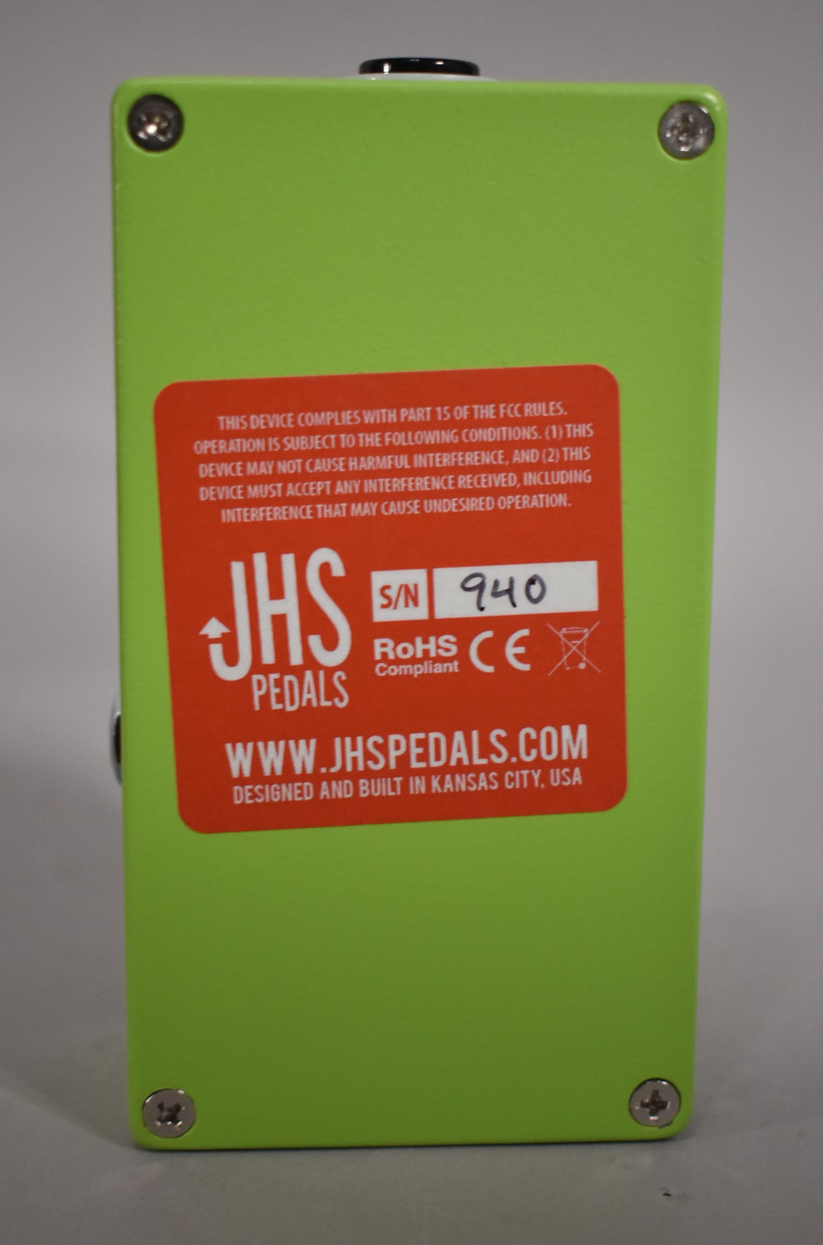 The bright green pedal is viewed from the back, revealing an orange sticker with product information and a handwritten serial number.