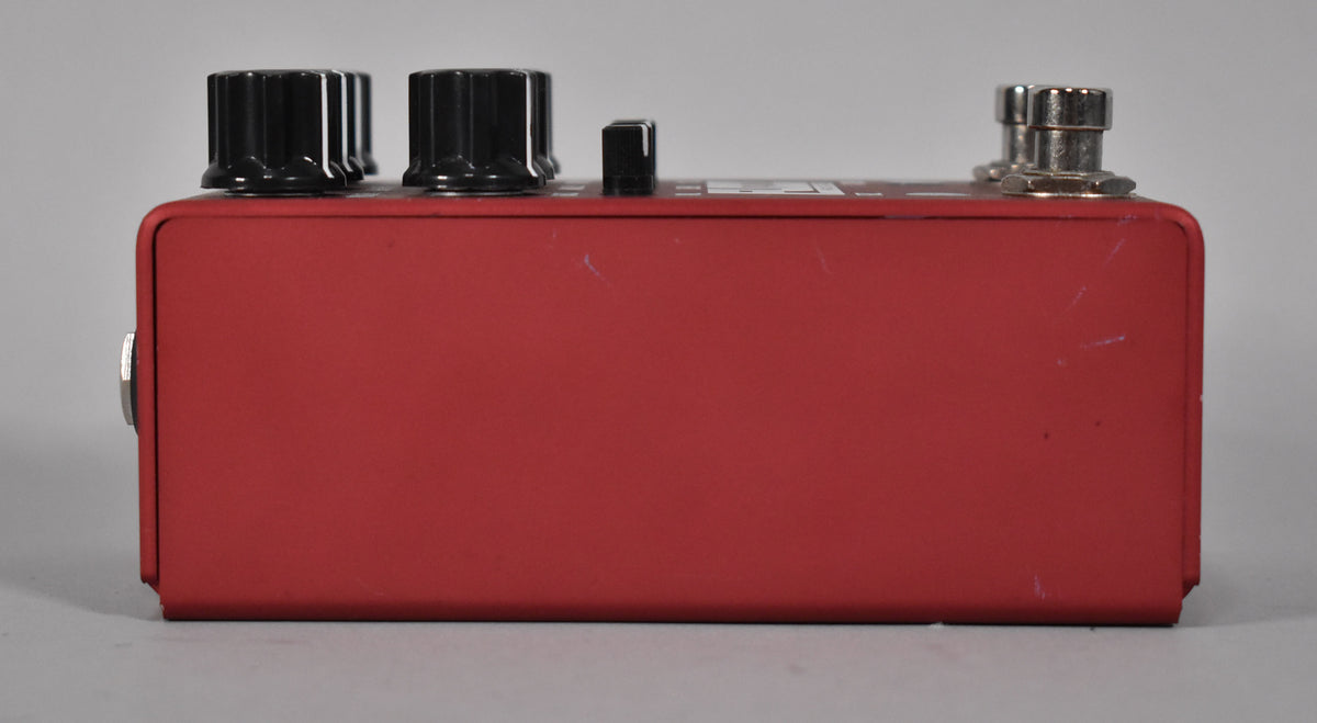 The red pedal is viewed from the left side, showing a silver input jack, three black knobs, a small black switch, and two silver jacks.