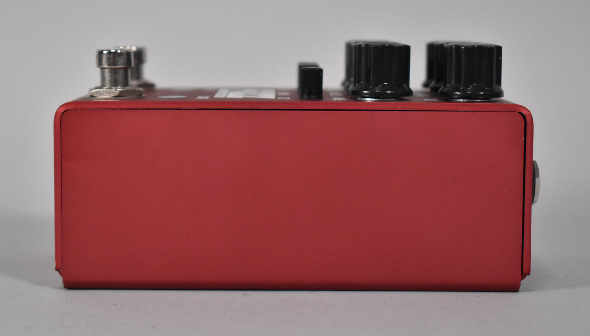 The red fuzz pedal is viewed from the right side, showing a few black control knobs and a silver jack on the top surface.