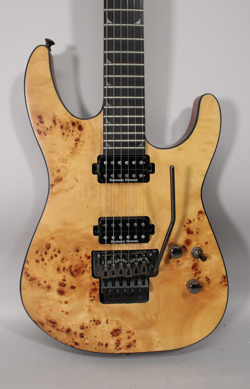 Full front view of a light tan electric guitar with dark brown burl wood grain, black humbucker pickups, and a Floyd Rose tremolo system.