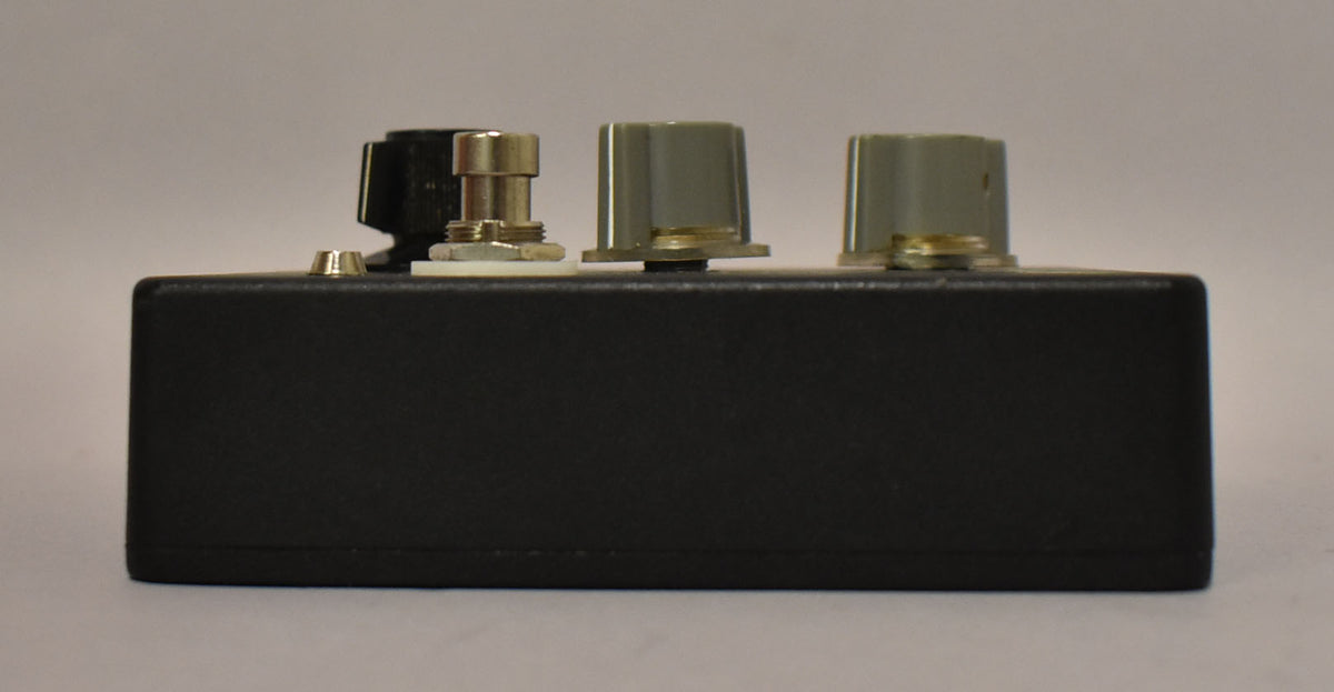 The black fuzz pedal is viewed from the side, showing a silver footswitch and two grey control knobs on top.