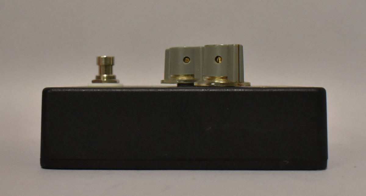 The black fuzz pedal is viewed from the side, showing a gold footswitch on the left and two gray control knobs on the right.