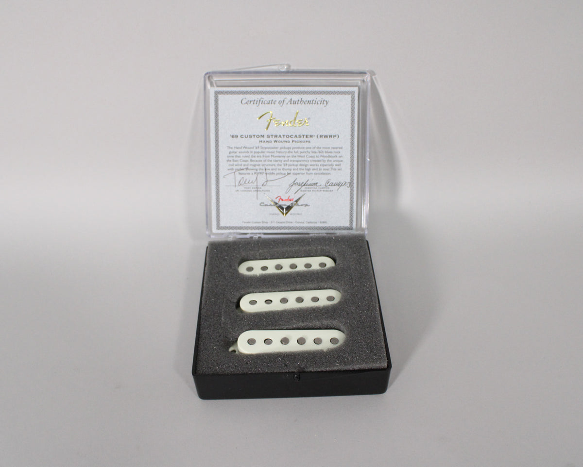 Three off-white single-coil pickups with exposed pole pieces are nestled in black foam, with a certificate of authenticity behind them.