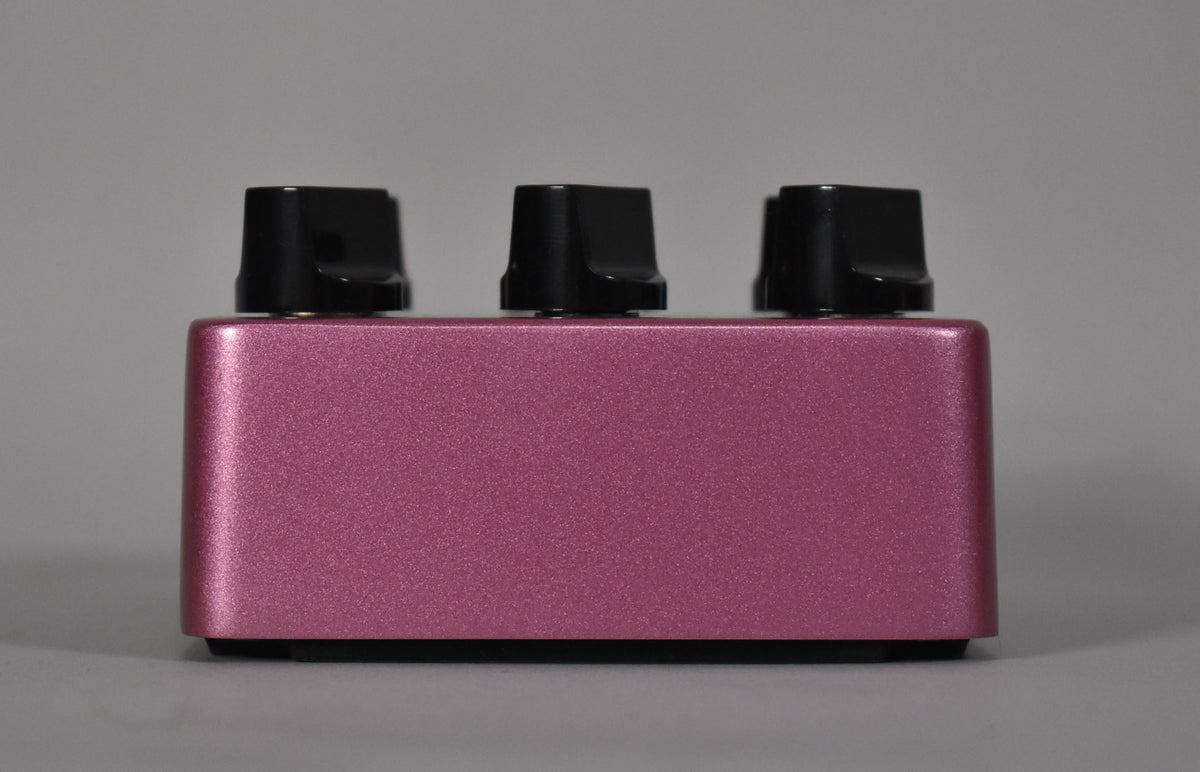 The fuchsia metallic pedal is viewed from the side, revealing three black control knobs along the top edge.