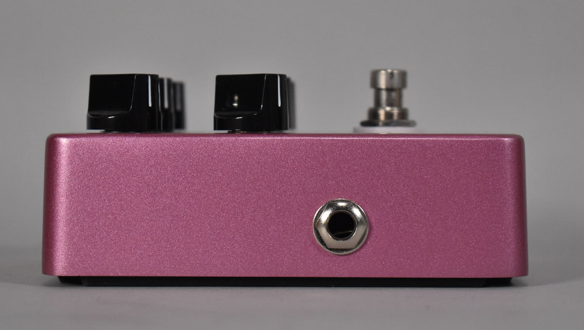 The fuchsia pedal is viewed from the side, showing a silver input jack, three black control knobs, and a silver footswitch.