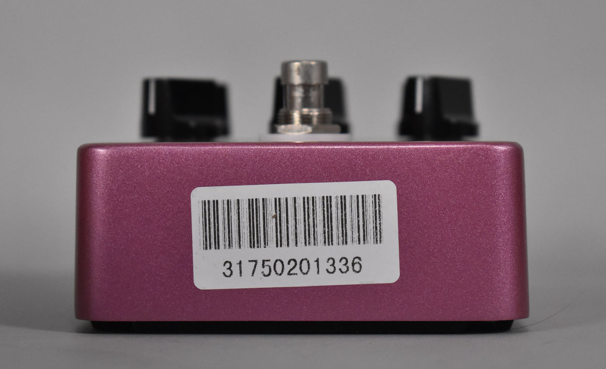 The fuchsia pedal is viewed from the back, showing a white barcode sticker on the metallic finish, with three black knobs visible above.