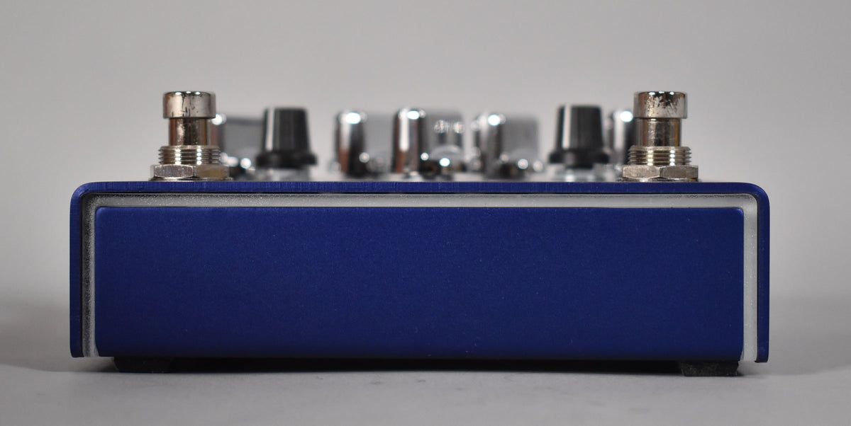 Side view of a blue guitar pedal with two silver footswitches and several chrome knobs on top.