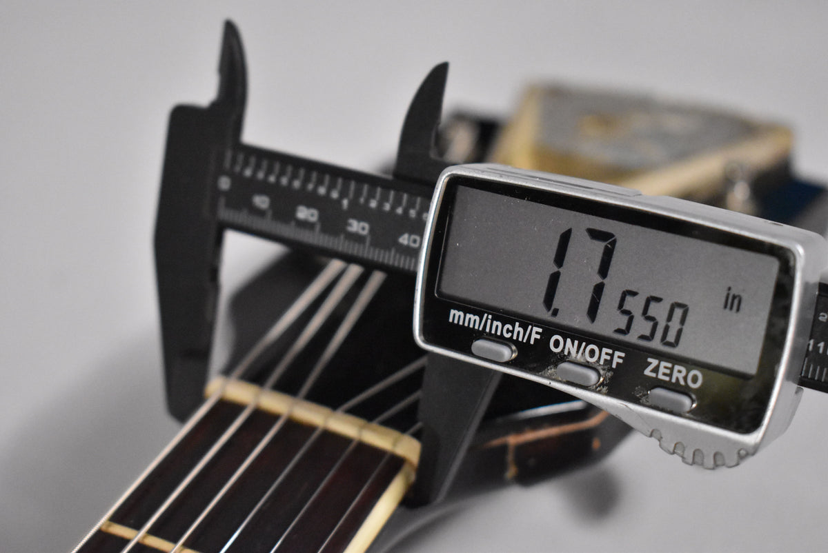 A digital caliper measures the guitar's nut width at 1.7550 inches, with the dark fretboard and light nut visible.