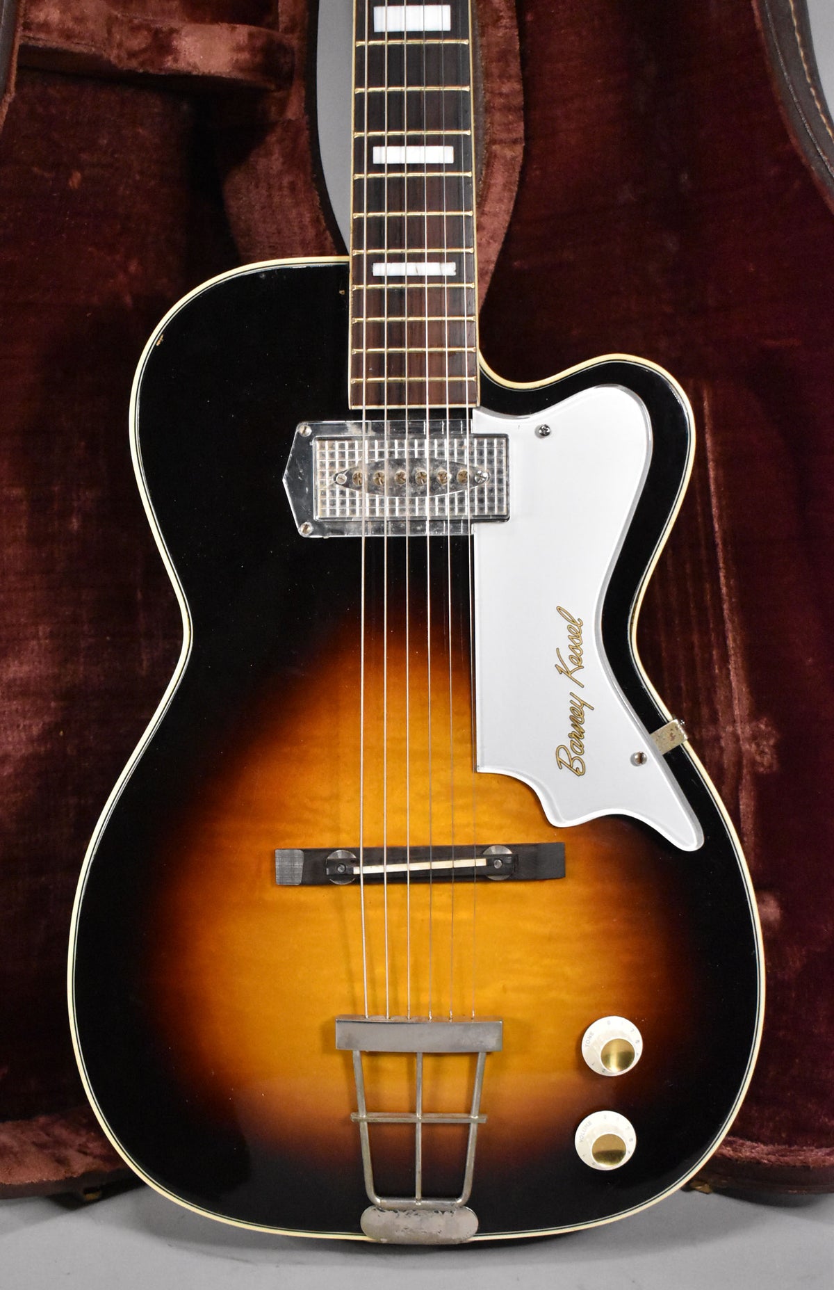The sunburst archtop guitar is angled slightly right, showing its white pickguard with gold lettering and two white control knobs.