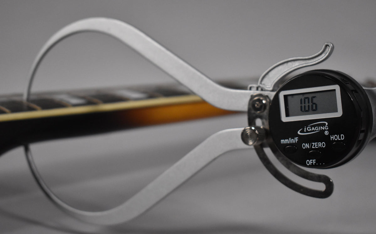 A digital caliper measures the sunburst neck's thickness at the first fret, displaying "1.06" on its screen.