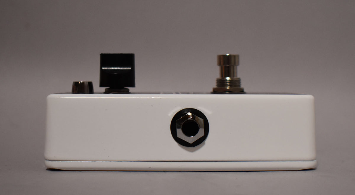 The white pedal is viewed from the side, showing a black input jack, a small black knob, a larger black knob, and a chrome footswitch.