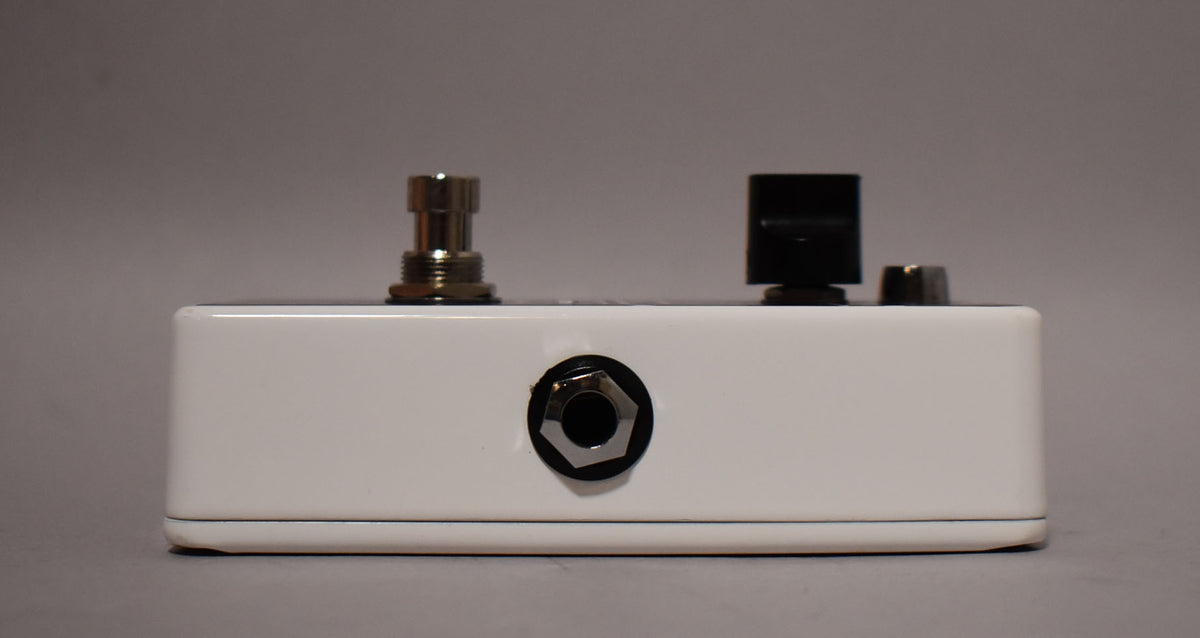 The white pedal is viewed from the side, showing a chrome input jack, a silver footswitch, and a black control knob.