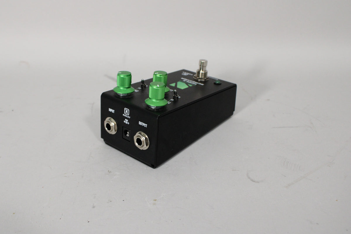 Black pedal with three green knobs, a silver footswitch, and input/output jacks, viewed from the front-left.