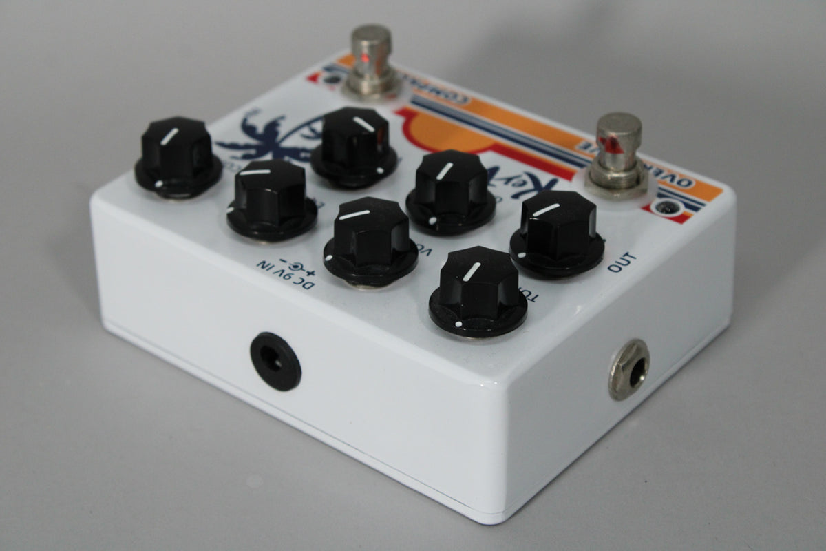 White pedal with a tropical graphic, six black control knobs, and two silver footswitches, viewed from the front-right.