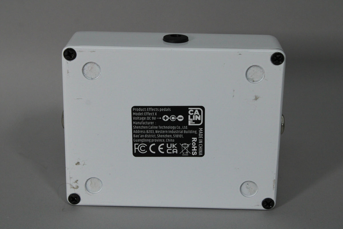 The white pedal is viewed from the bottom, showing four black screws, four circular rubber feet, and a black label with white text.