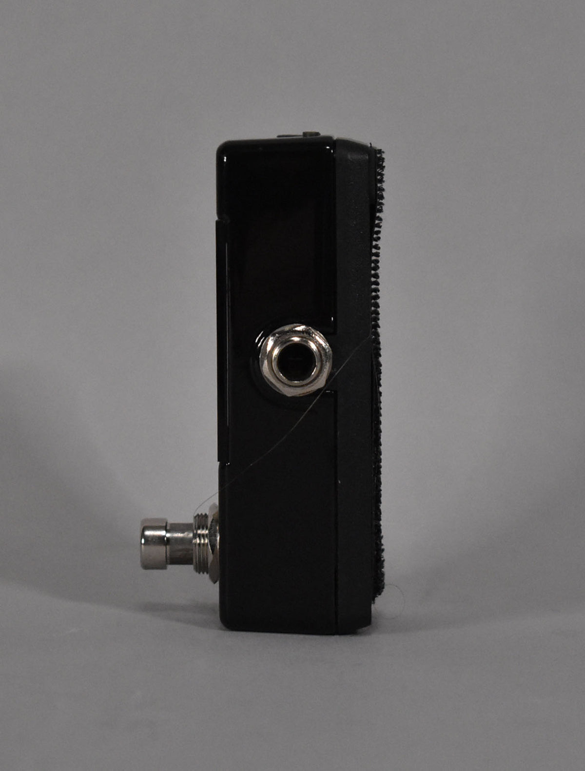 The black mini tuner pedal is viewed from the side, showing a silver input jack and a threaded silver footswitch shaft.