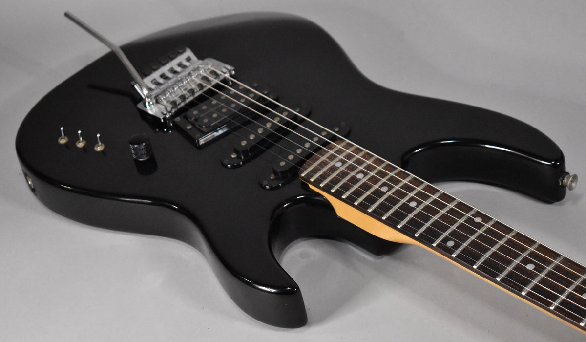 A black electric guitar with a maple neck and rosewood fretboard, showing the body, bridge, pickups, controls, and part of the neck.