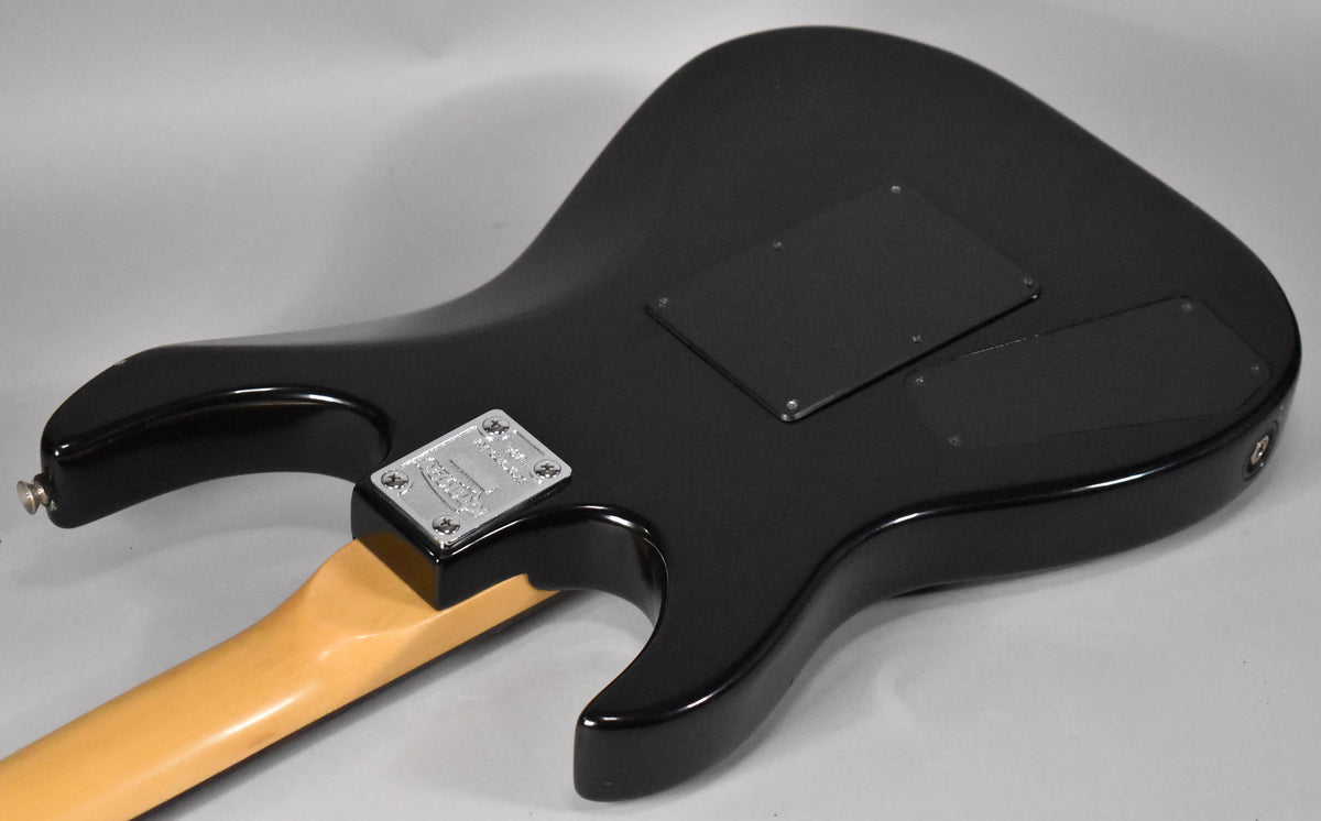 The back of a black electric guitar with a glossy finish, showing a light wood neck and two black control cavity covers.