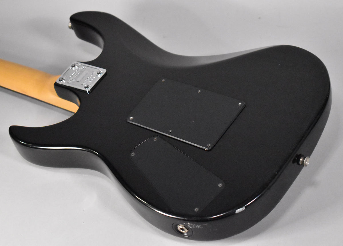 The black electric guitar's back is angled, showing two control cavity covers, a chrome neck plate, and a worn output jack.